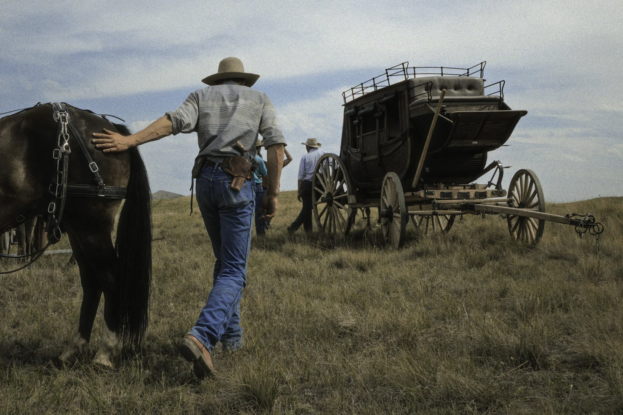A cowboy walks beside his horse across open grassland, guiding it toward a waiting stagecoach. The scene captures a quiet moment of daily life rooted in tradition, work, and connection to the land.
