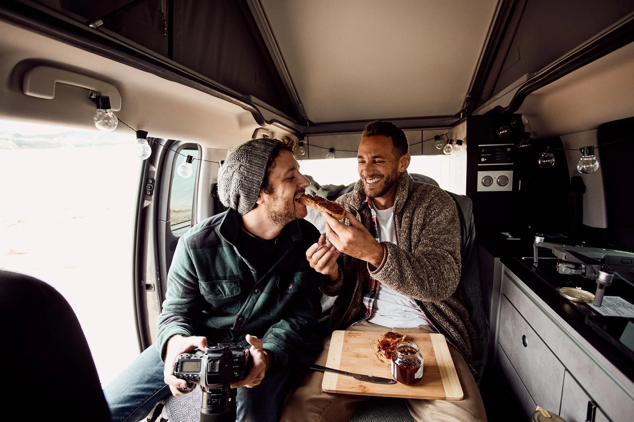 Two men enjoying a meal inside a camper van, one offering the other a slice of bread with jam, with kitchen appliances and string lights visible in the background.