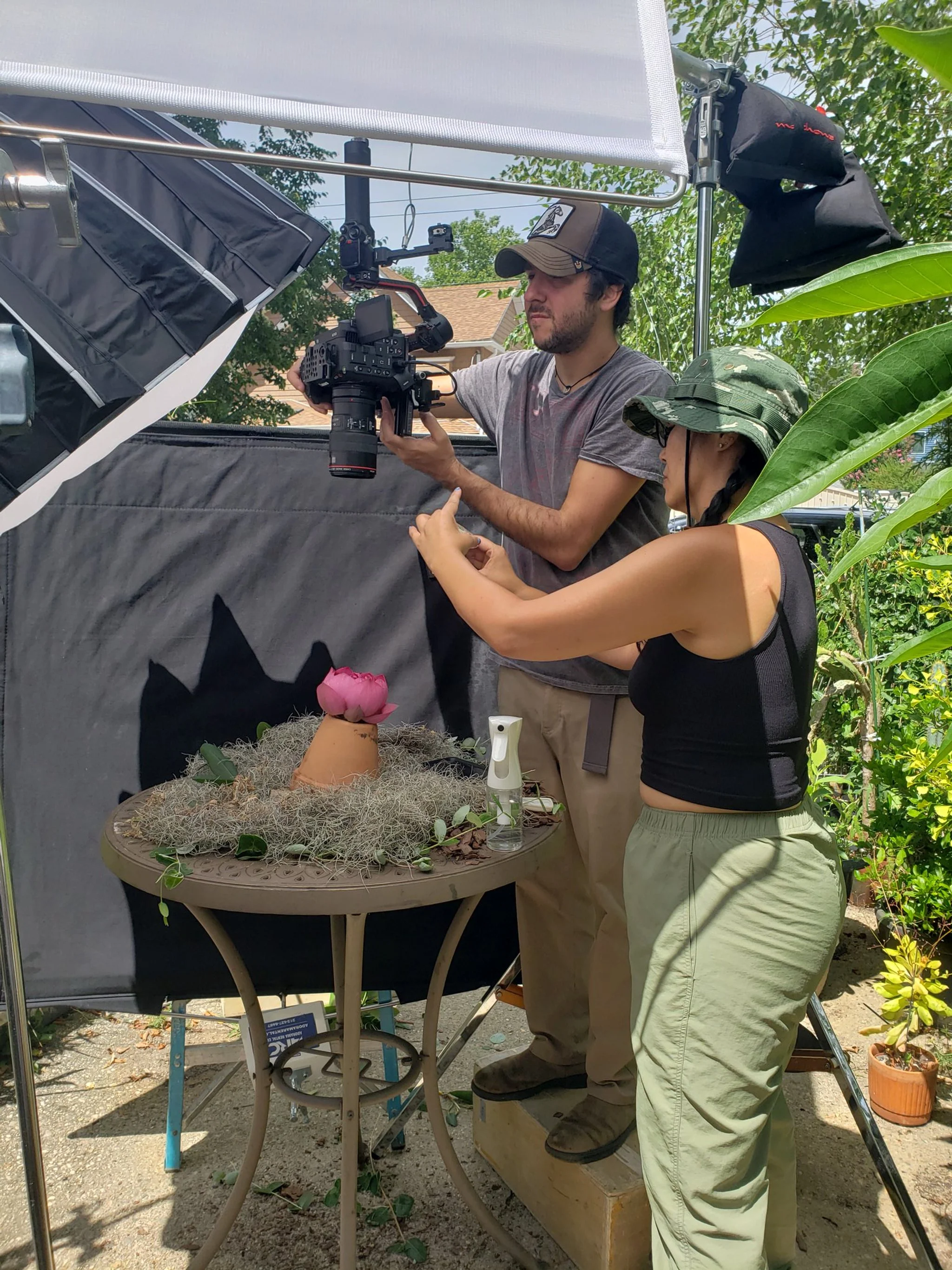 Two people working on a photo shoot outdoors under a light tent, with a terracotta pot with a pink flower on a table covered with moss and plants, surrounded by greenery.
