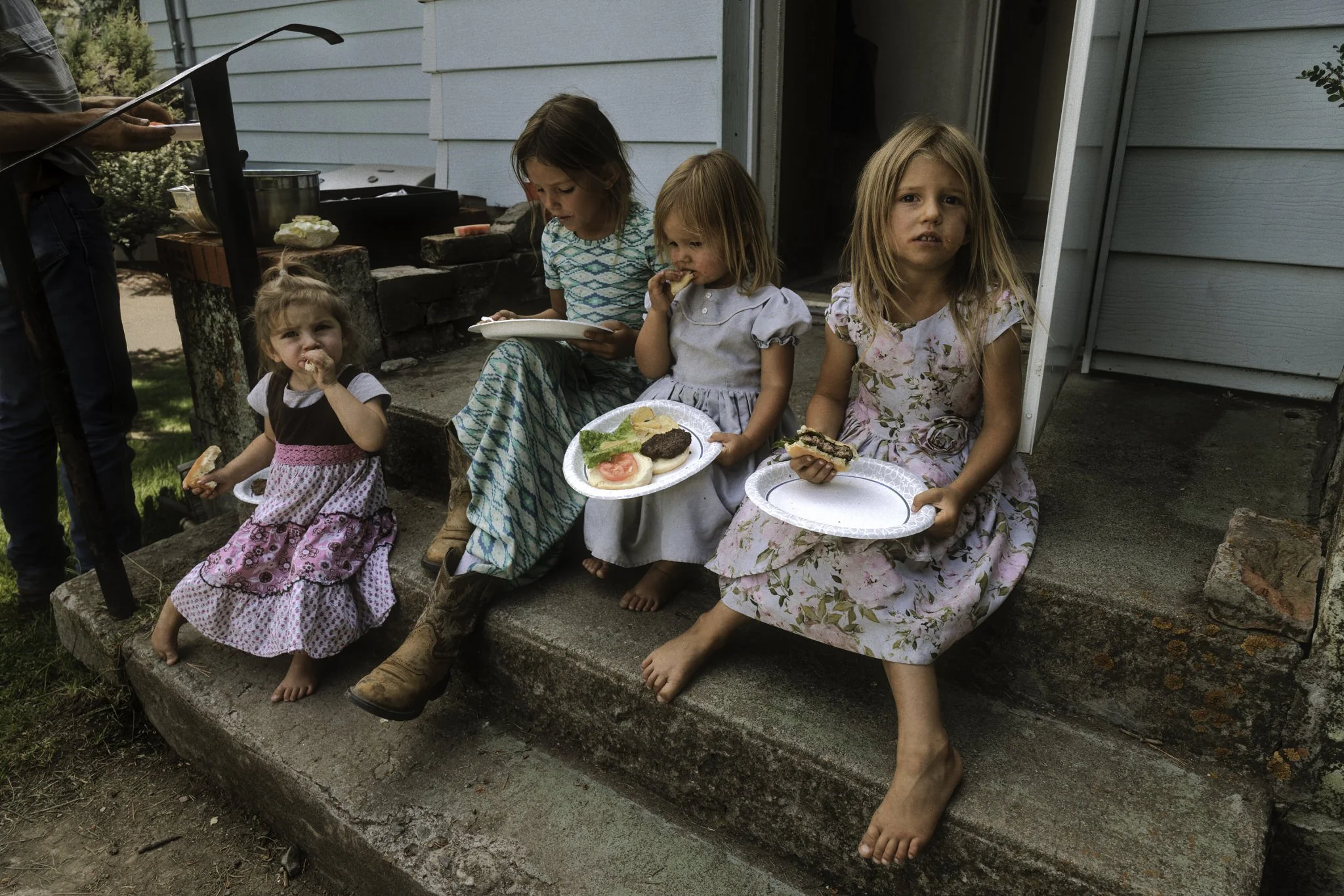 Four young girls sit on the front steps of a house, eating together on paper plates. The moment captures a simple slice of rural family life, shaped by home, routine, and togetherness.
