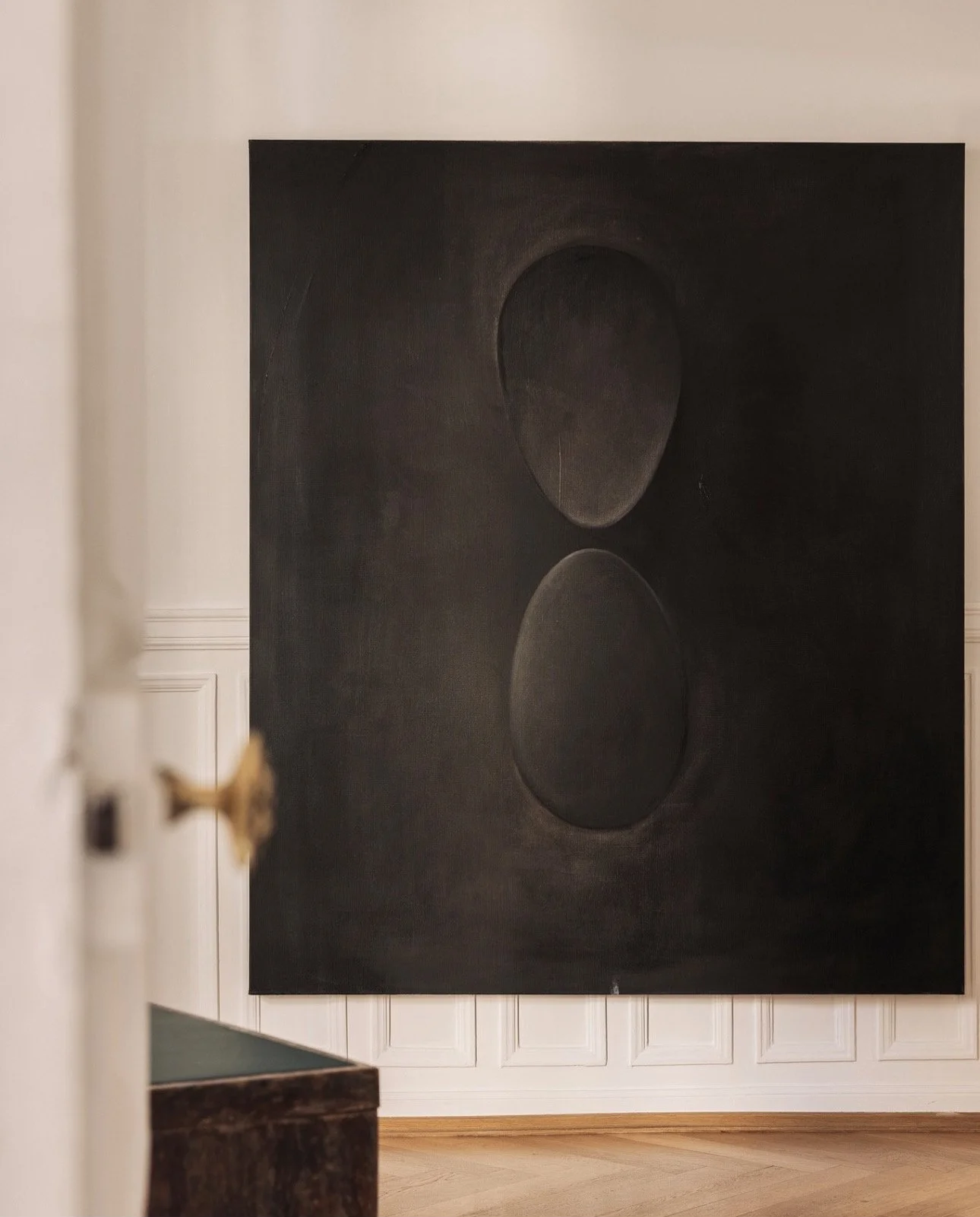 Large black painting featuring two circular shapes, one above the other, with subtle shading and reflections, displayed on a white wall in an art gallery.