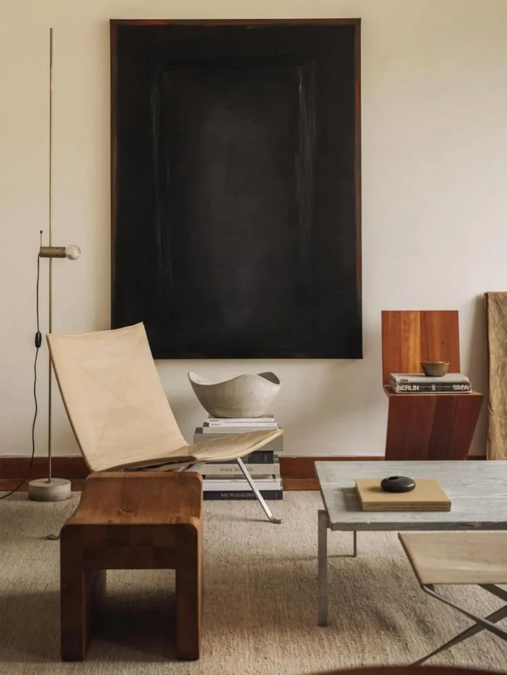 A living room corner featuring a beige armchair, a wooden side table, a stack of books with a ceramic bowl on top, a gray marble coffee table with a book and a small decorative object, and a black chalkboard wall with a white wall lamp beside it.