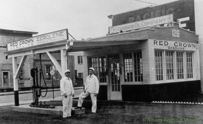 Two men in white uniforms stand beside the single pump at the Red Crown Gas Station, Second & Ellsworth, c. 1912. The Albany Stables can been seen at the far left in this photo.