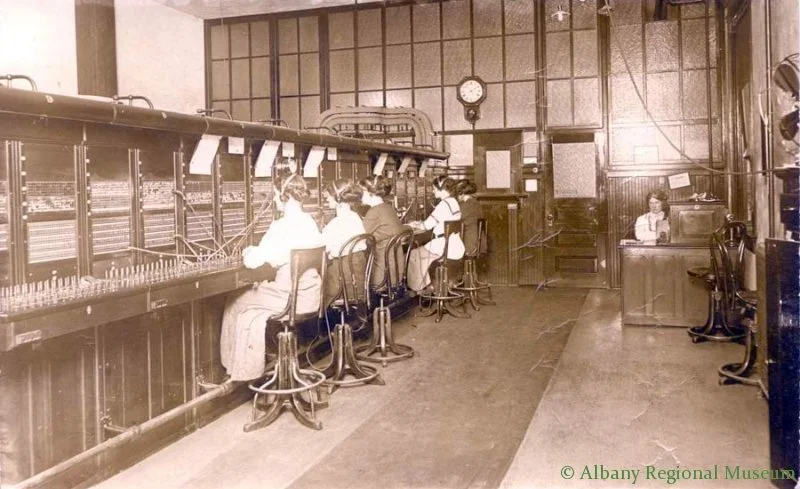 Photographic Print on Postal card of Albany Bell telephone office when it first moved into the Burggraf Building on 2nd Ave in the 100 West block. Shows 5 women at telephone switchboard and 1 woman, Maybe the supervisor, seated at desk facing camera. Has names on back but are hard to read. c. 1900.