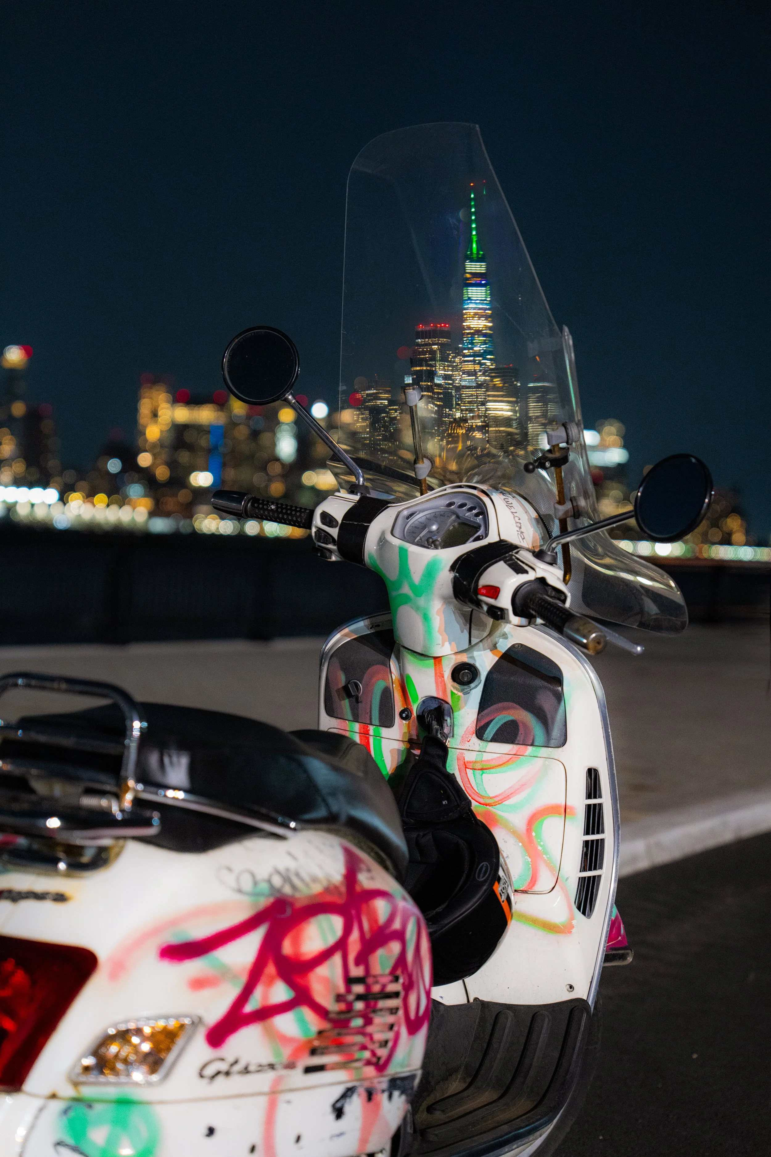 Vibrant night shot of a custom graffiti-covered scooter overlooking the illuminated NYC skyline and Empire State Building. Iconic urban photography.
