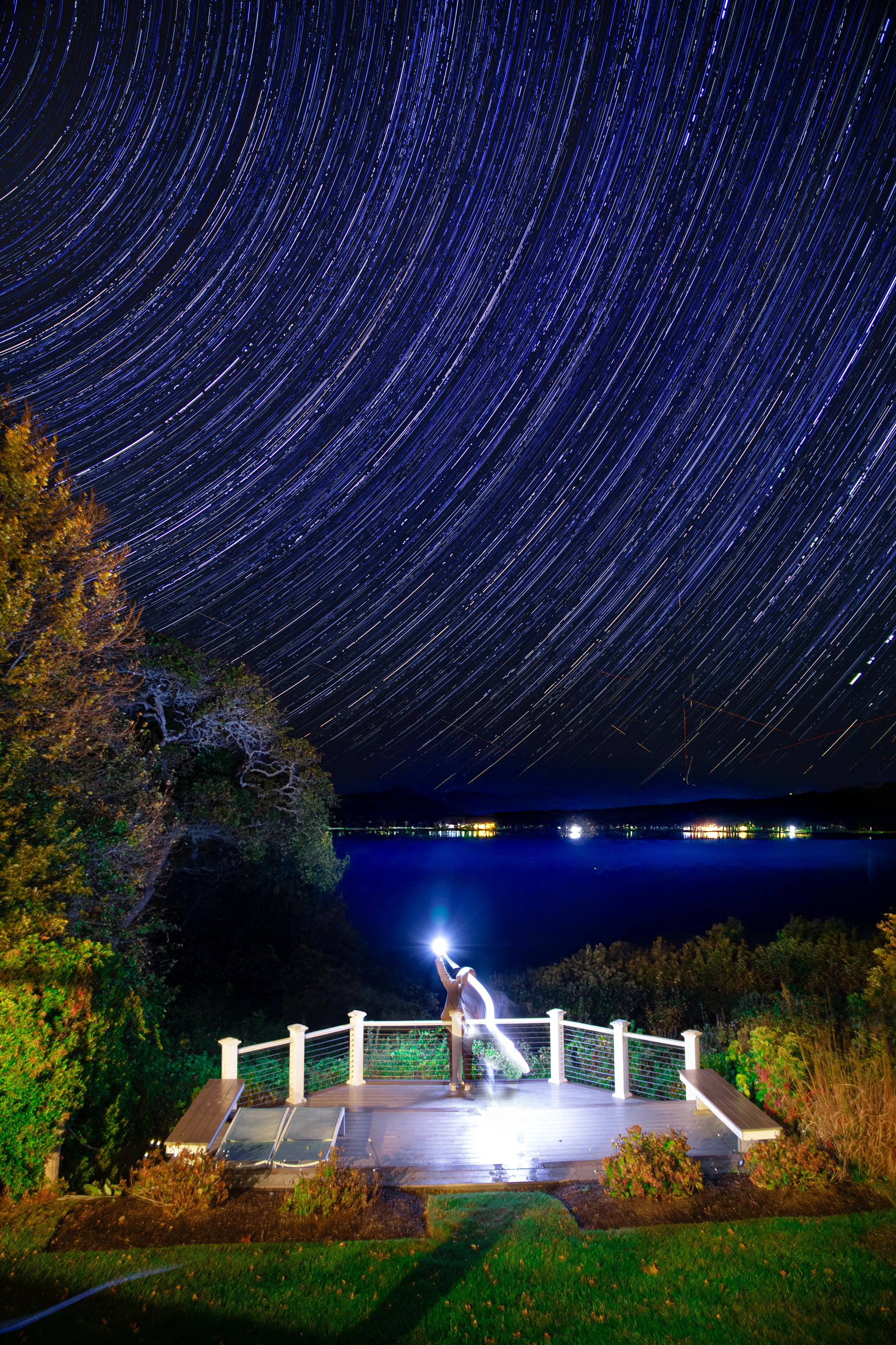 Mesmerizing long exposure night shot showing swirling blue star trails in the sky over a brightly lit outdoor deck and scenic waterfront landscape.