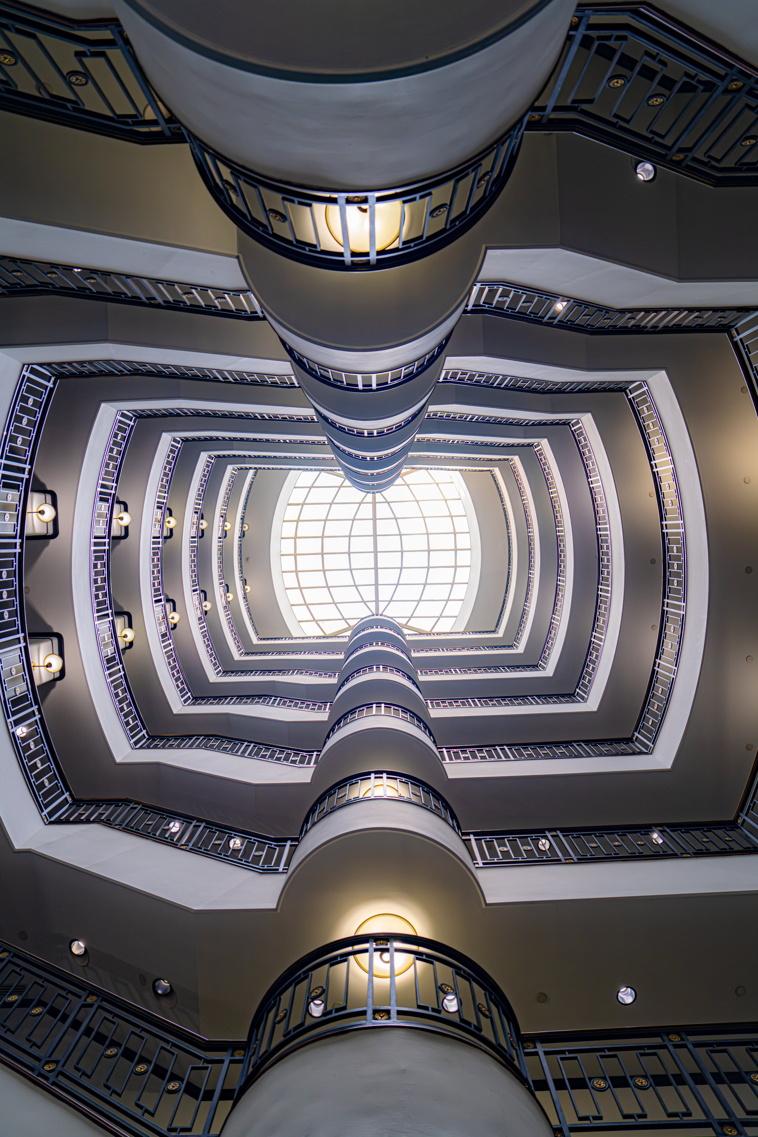 Stunning low-angle shot looking up through a multi-story spiral staircase toward a glowing skylight dome. Perfect for modern architecture, interior design, and urban photography lovers.