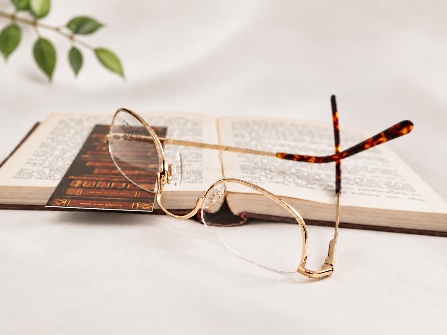 B-AQ gold half-eye reading glasses resting on an open book, with softly blurred green leaves in the background