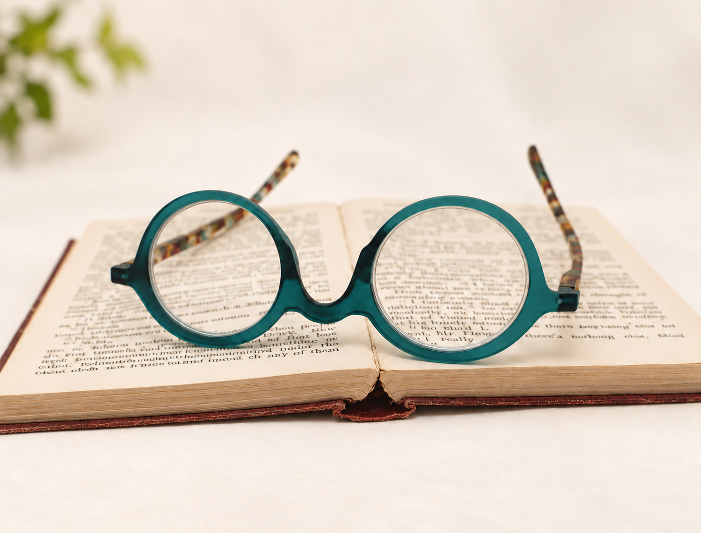 Front view of Ikaly Lisboa teal and tortoiseshell-patterned reading glasses resting upside down on an open book