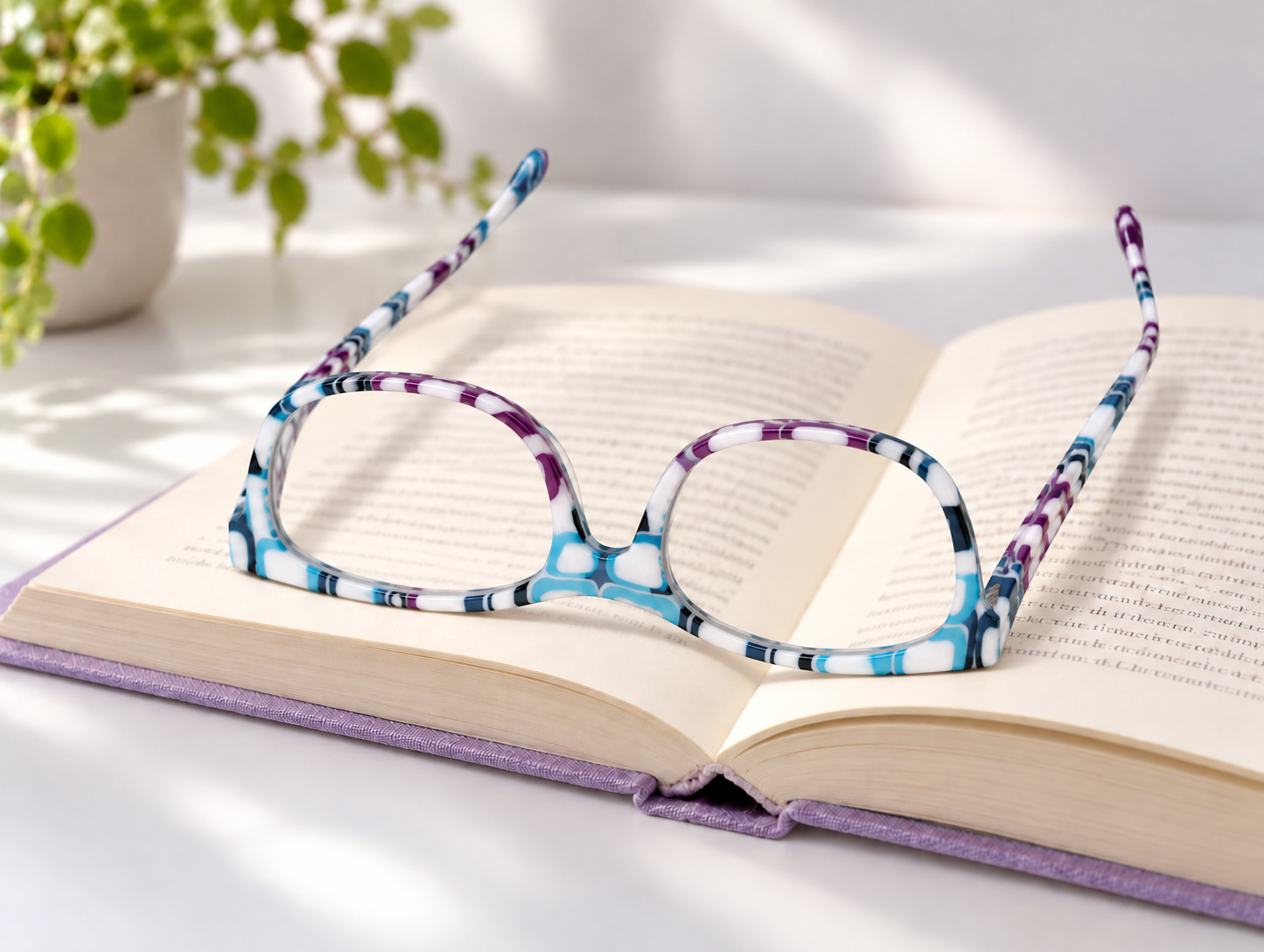 L56 white, blue and purple patterned reading glasses resting upside down on an open lavender-covered book on a white desk beside a plant.