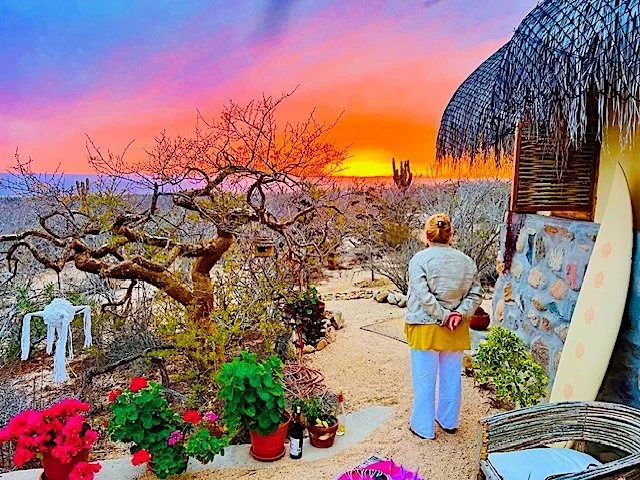 A woman standing outside near a stone building at sunset, with a beach and cactus in the distance, surrounded by potted plants and flowers.