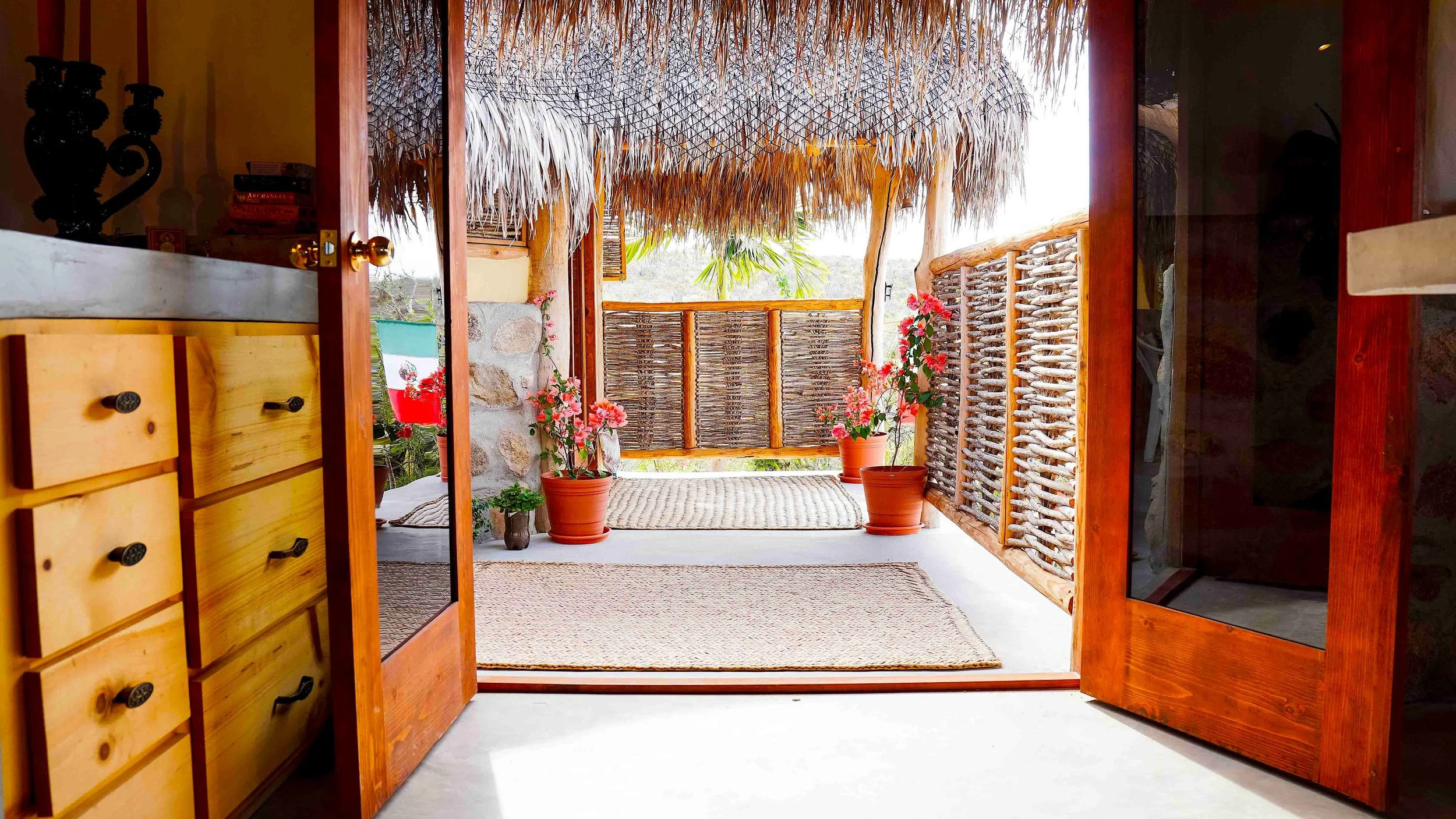 View from inside a room with open wooden doors looking out to a balcony with potted pink flowers, a bamboo fence, and a thatched roof roof overhang.