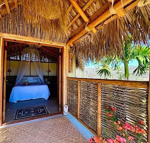 View of a thatched-roof balcony with a bed inside a tropical bungalow, overlooking lush green trees and pink flowers.