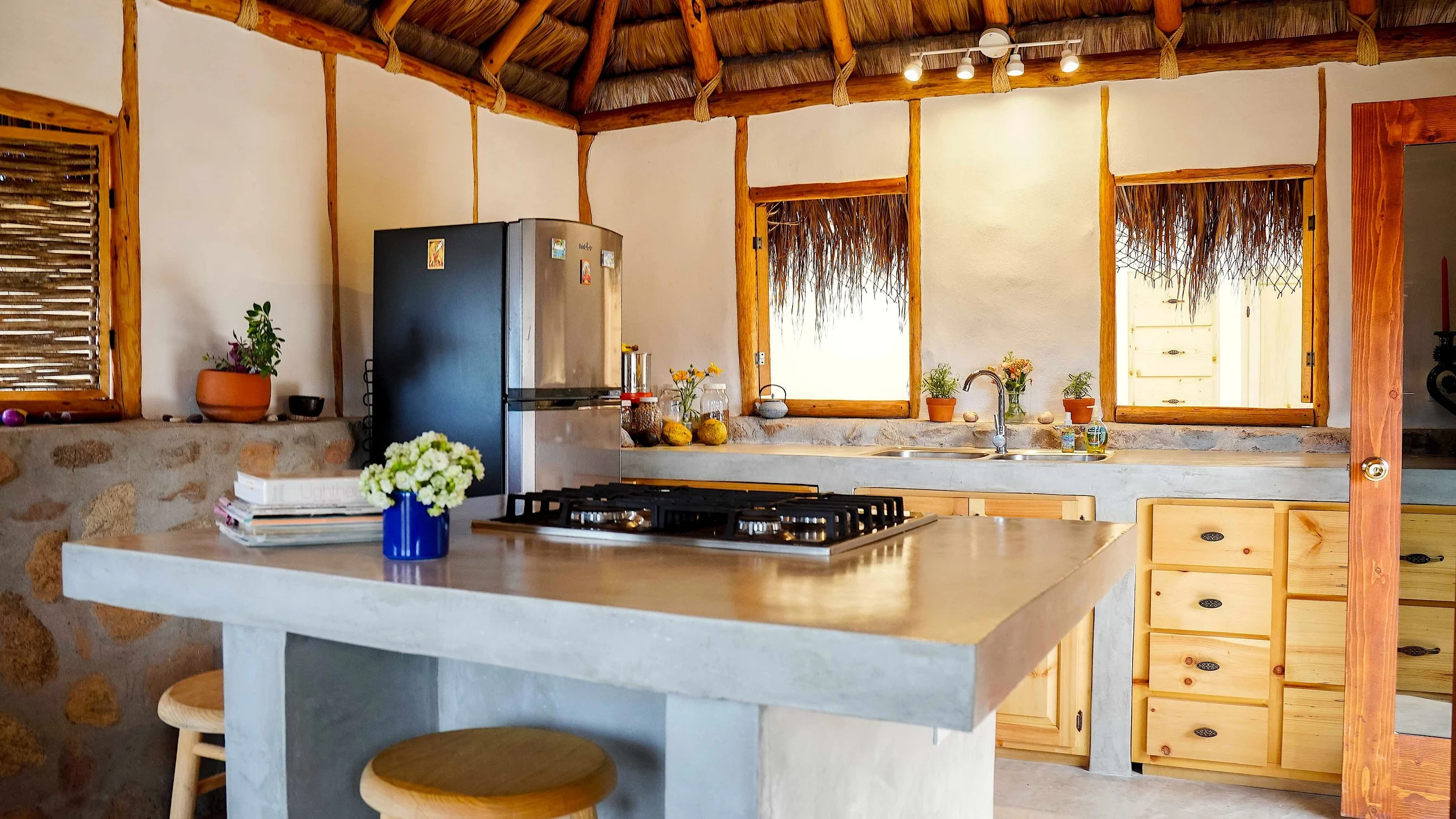 A rustic kitchen with stone walls, wooden cabinets, and a thatched ceiling. There are two windows with bamboo blinds, a black refrigerator, and a wooden countertop with a sink. Decor includes potted plants, flowers, and small kitchen items.