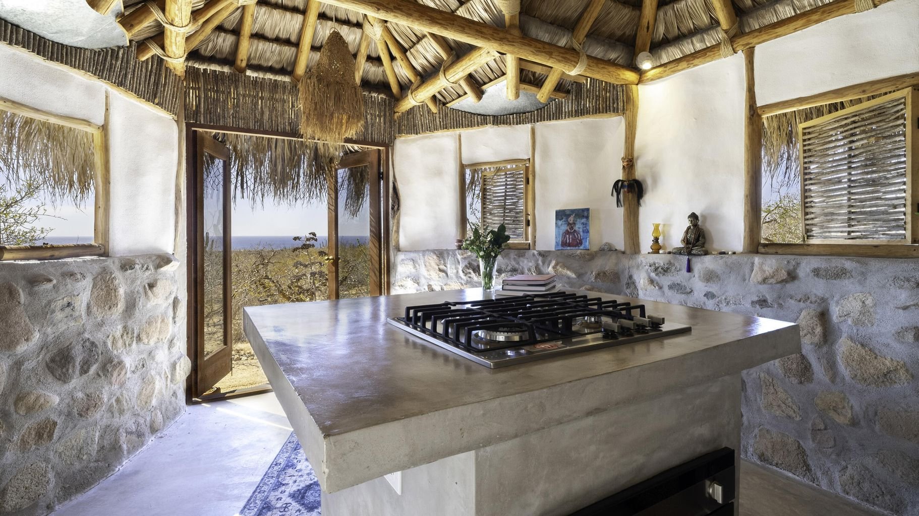 Interior of a rustic cottage with stone walls and thatched roof, featuring a kitchen island with a built-in gas stove, and windows showing an outdoor landscape with trees and distant ocean.