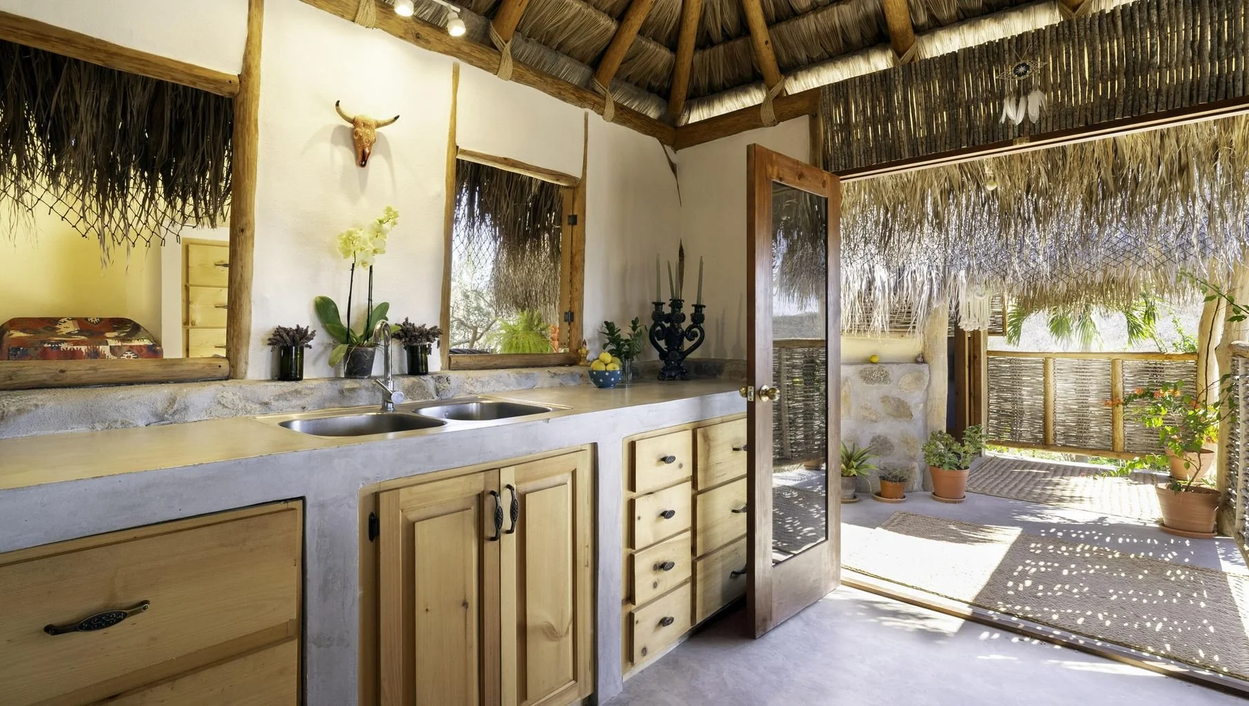 Interior of a thatched-roof kitchen with wooden cabinets, a double sink, and open doors leading to an outdoor patio with potted plants and wicker furniture.