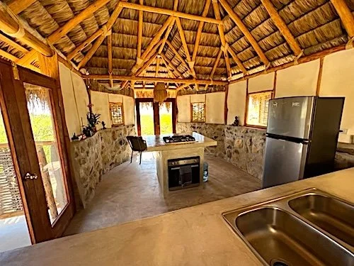 Interior of a circular, rustic hut with a thatched roof, stone wall accents, a small kitchen area with a double sink, and a refrigerator.
