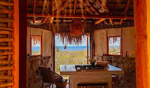 Interior of a rustic beach hut with a view of the ocean, showing a wooden table, chairs, and straw accents.