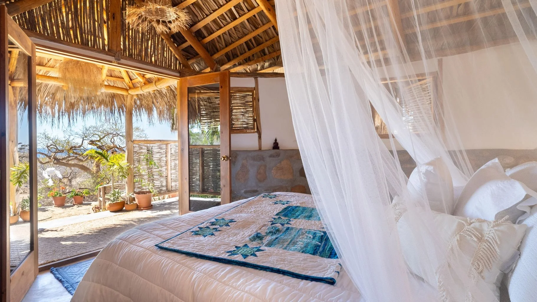 Bedroom with a mosquito net hanging over a bed, open wooden door leading to a sunny outdoor terrace with potted plants, thatched roof, and view of trees outside.