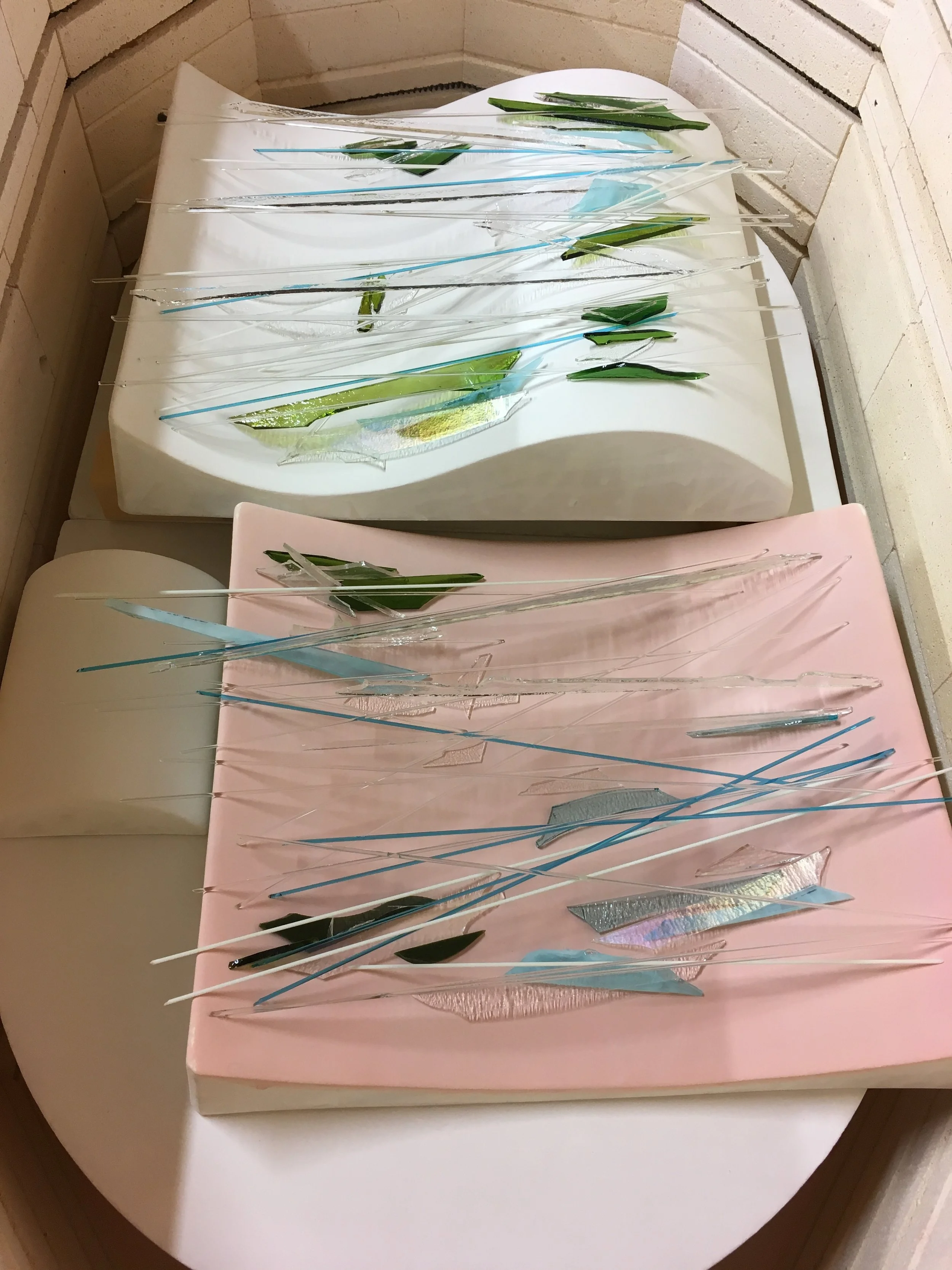 Finally, here the strips of sheet glass and stringers placed over the molds — to be slumped into shapes that will (later) be combined with more glass and fired much hotter. (BEFORE firing.)