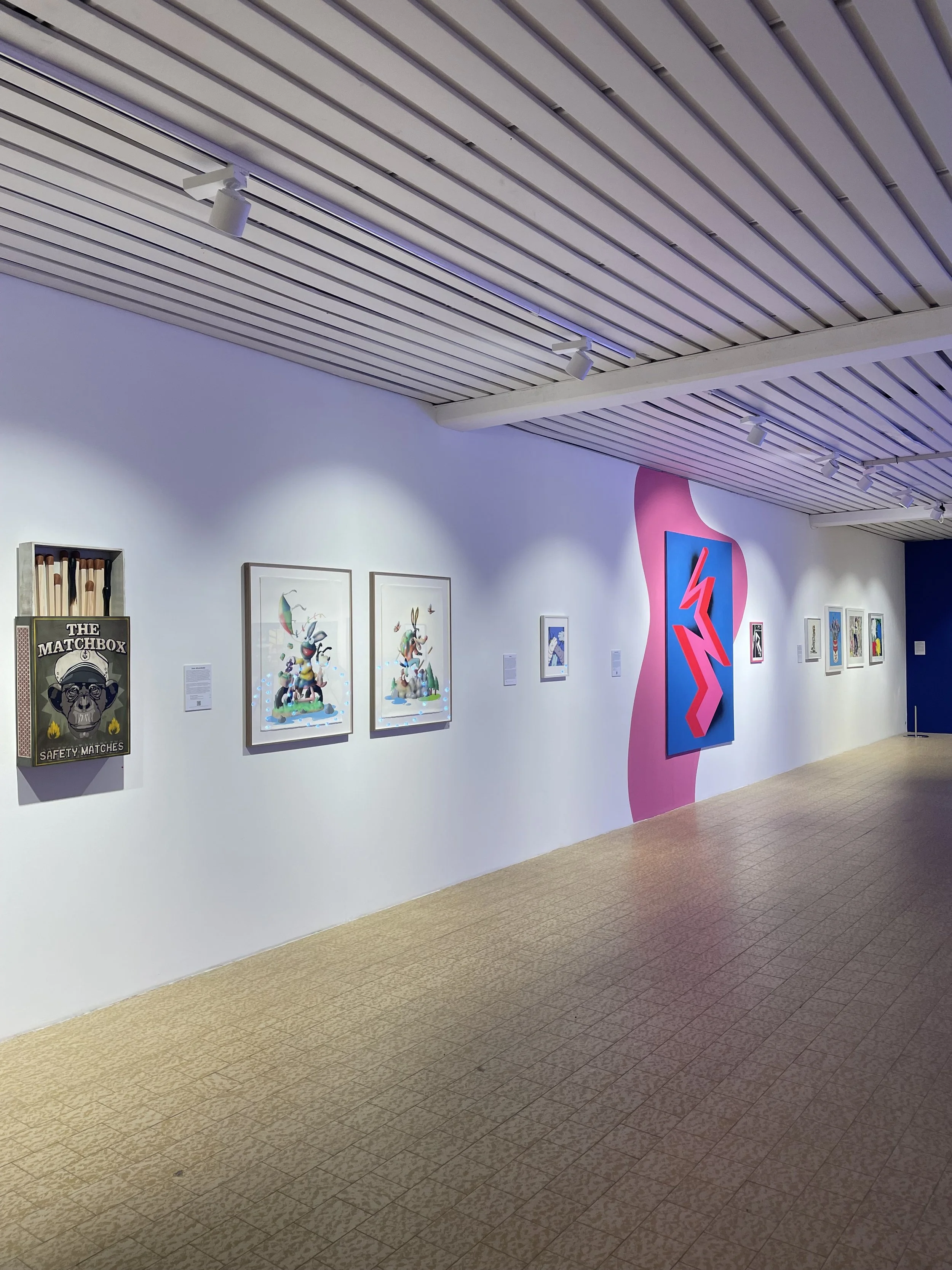 An art gallery wall displaying various framed artworks, with a pink and blue abstract sculpture on the right, and a safety matches display in the left corner. The gallery has a wooden slat ceiling with adjustable lighting.
