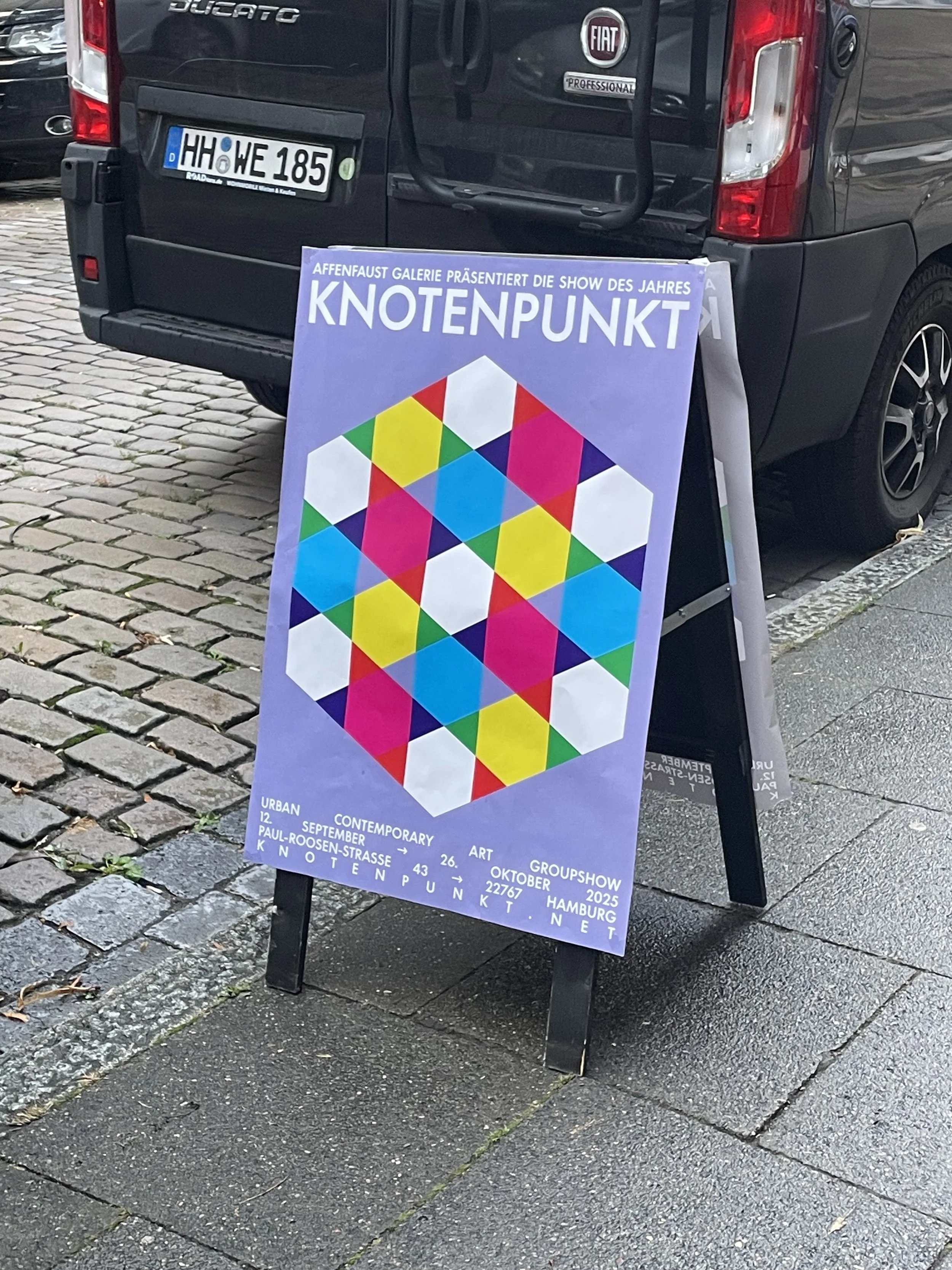 A black van with European license plates parked on a cobblestone street. A colorful signboard in front advertises an art group show titled "KNOTENPUNKT" at the Affenfaust Gallery in Hamburg, Germany, from September 12 to October 26, 2025.