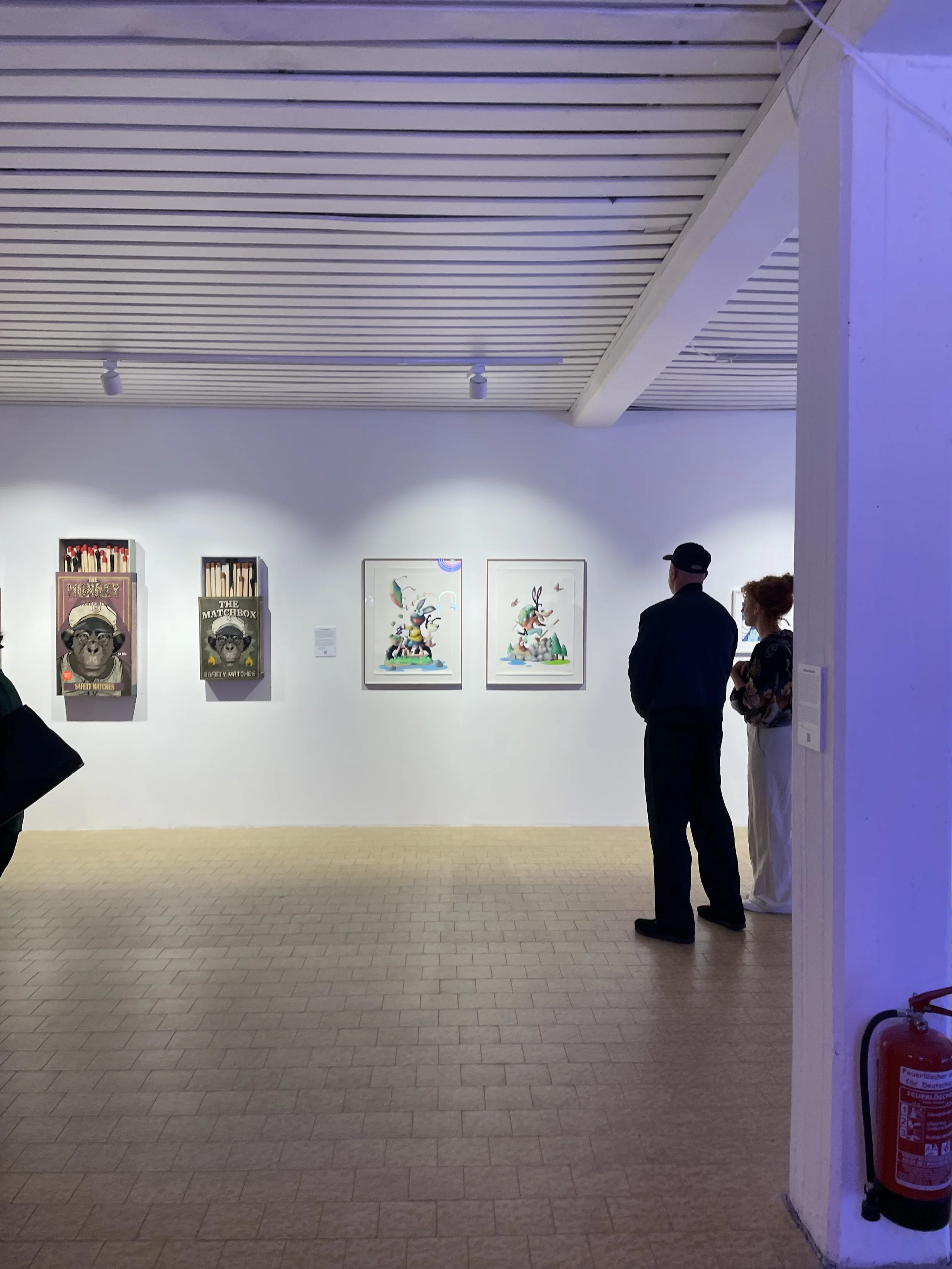 People standing in an art gallery viewing paintings on the wall, with a fire extinguisher placed nearby.