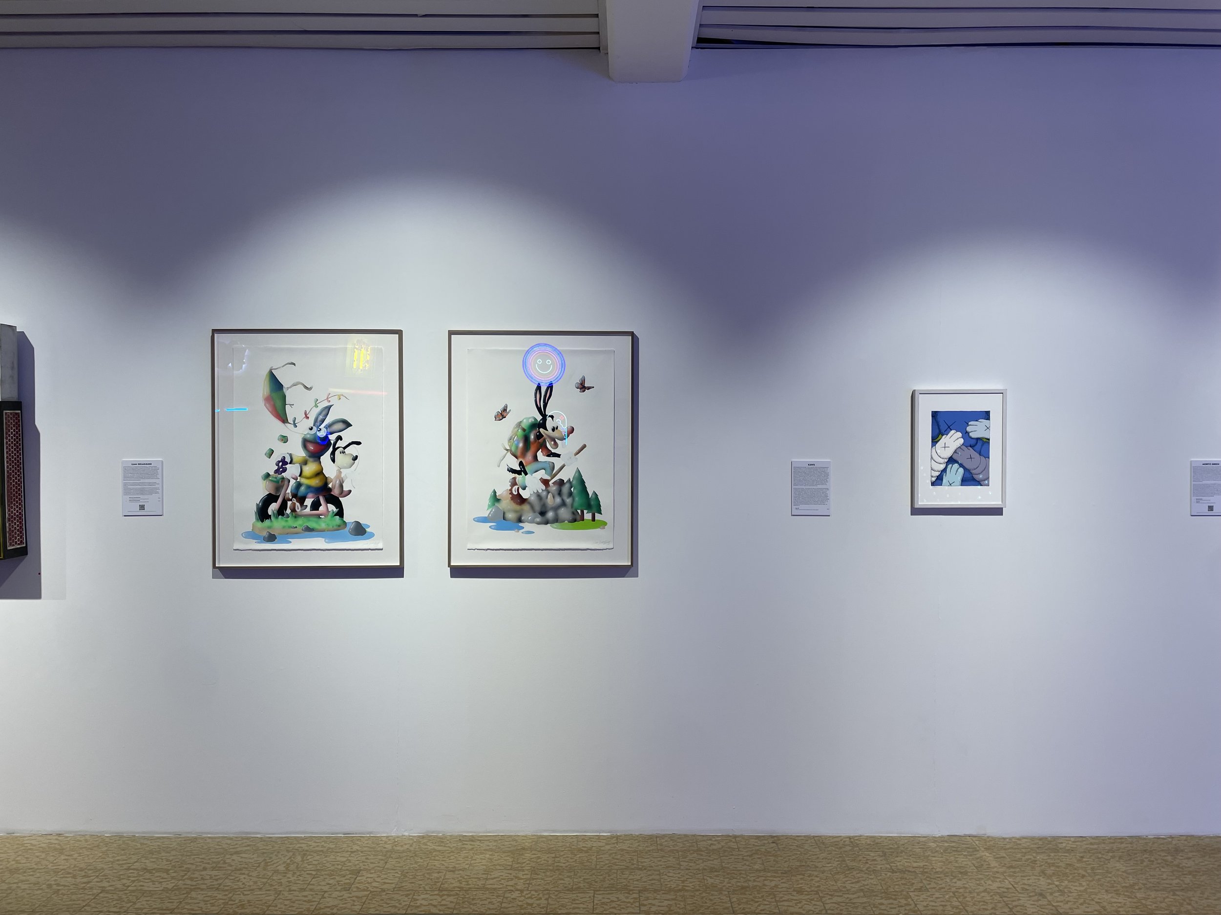 Three framed artworks on a white gallery wall, with descriptions underneath each, and a carpeted floor in the foreground.