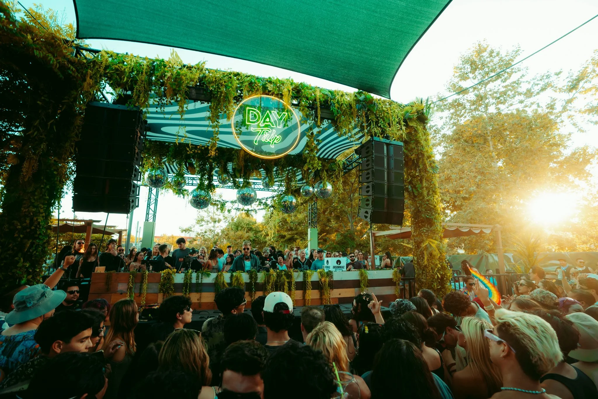 Crowd of people at an outdoor music festival during sunset, with a DJ performing on a stage decorated with greenery and a neon sign that reads 'Day Trip'.