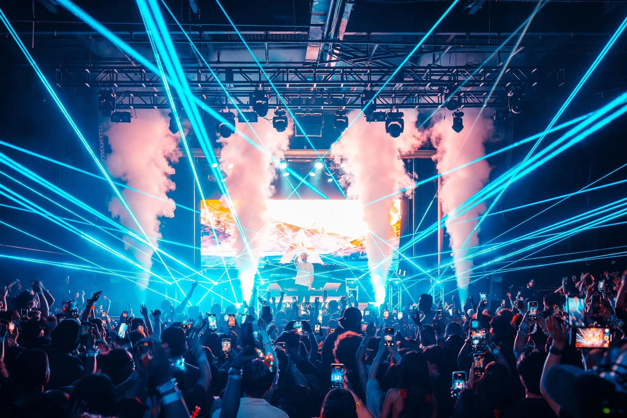 A live concert with a DJ on stage, colorful laser lights, smoke effects, and a large crowd of attendees taking photos and videos.