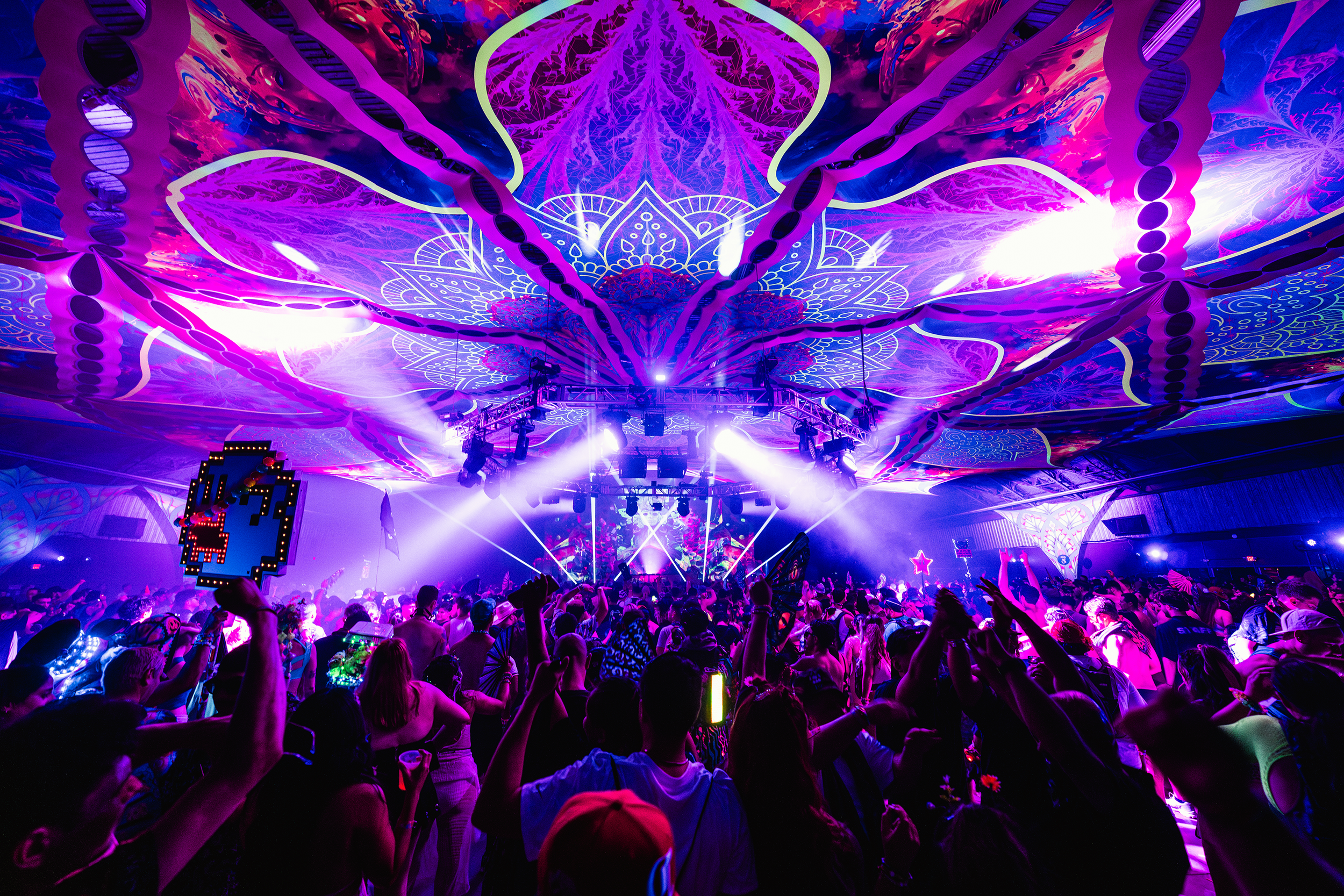 A vibrant nightclub scene with a crowd dancing under colorful, psychedelic ceiling lights, neon decorations, and stage lighting.
