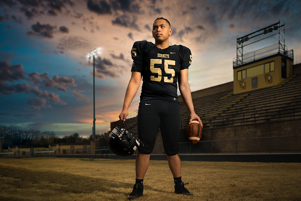 a high school senior wearing a black uniform stands on the field at sunset  holding a helmet in one hand and a football in the other by Don Evans Photography in Greensboro NC.jpg