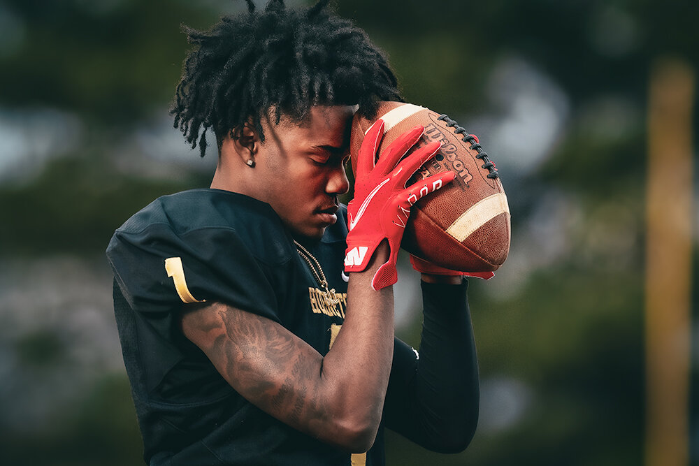 Fooball player wearing red gloves stands on the field and holds the ball to his forehead in thought during a senior portrait from Don Evans Photography in Greensboro NC.jpg