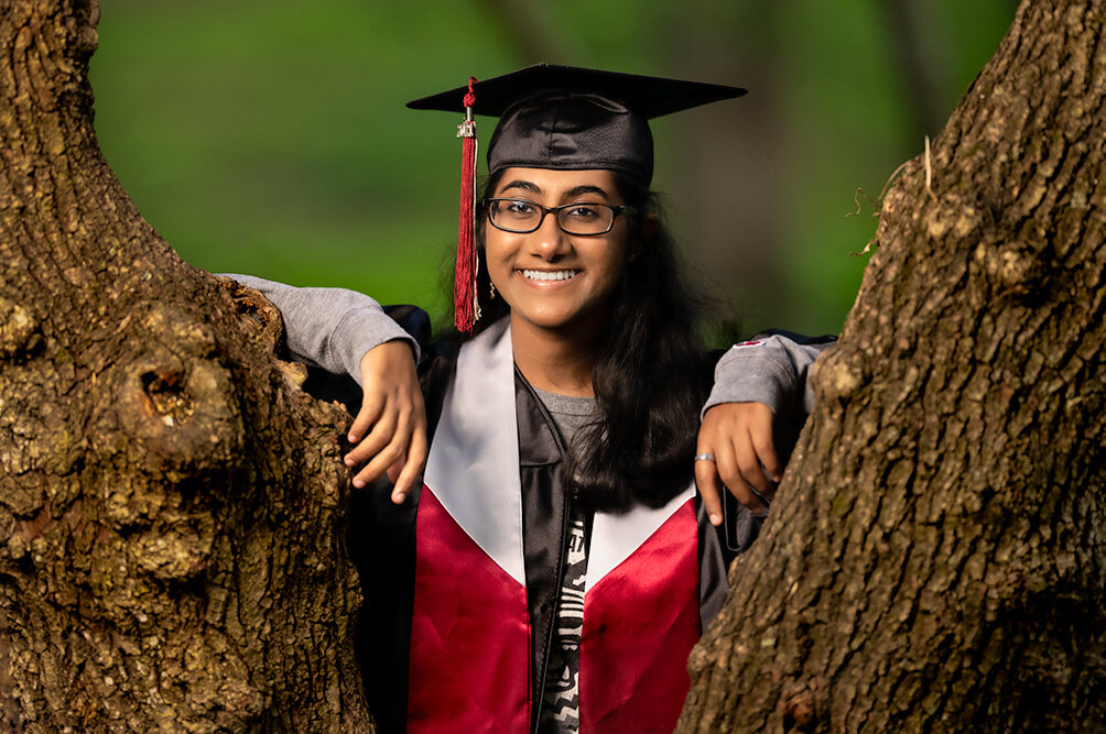 Early College at Guilford senior wearing her cap and gown leans up against two trees while posing during her senior pictures photo session with Don Evans Photography in Greensboro NC.jpg