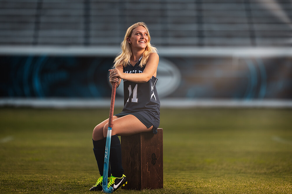 high school field hockey senior in blue uniform sits at mid-field in a stadium sunset during her senior sports pictures with Don Evans Photography in Greensboro NC.jpg