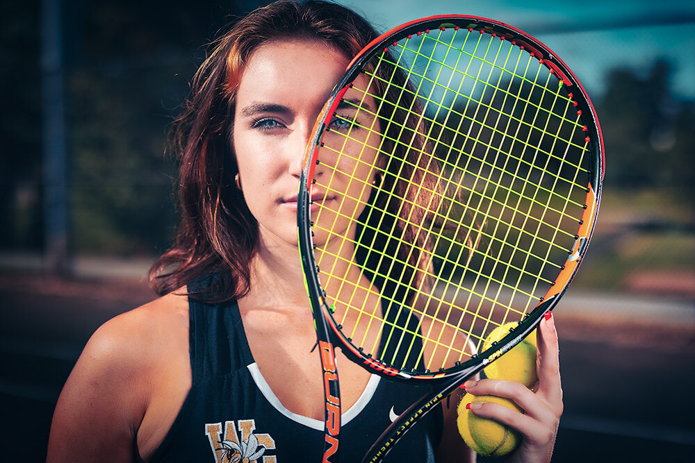 high school senior tennis player holds up her racket on the court while posing for her senior picture with Don Evans Photography in Greensboro NC.jpg