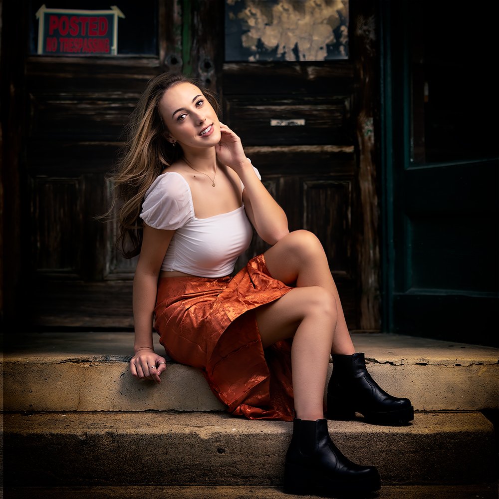 Don Evans Photography in Greensboro high school senior wearing an orange skirt and white top sits in front of an old building while posing for her Senior Pictures photo session.jpg