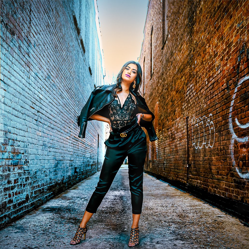 Don Evans Photography in Greensboro woman wearing black pants and top with a black leather jacket poses with hands on hips in an alley during her outdoor photo session