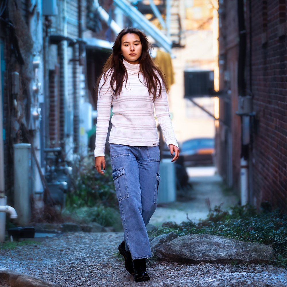 Don Evans Photography in Greensboro high school senior wearing jeans and white sweater walks down an alley during her senior pictures photo session