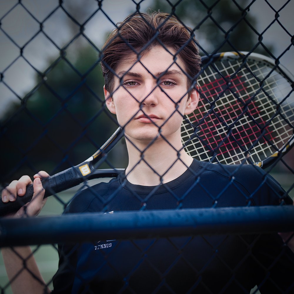 high school senior tennis player holds up his racket and leans against the court fence while posing for his senior picture with Don Evans Photography in Greensboro NC
