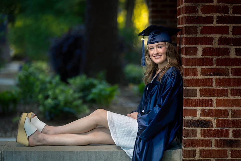 Don Evans Photography in Greensboro high school senior wearing a blue cap and gown sits on in an archway on campus while posing for her Senior Pictures photo session