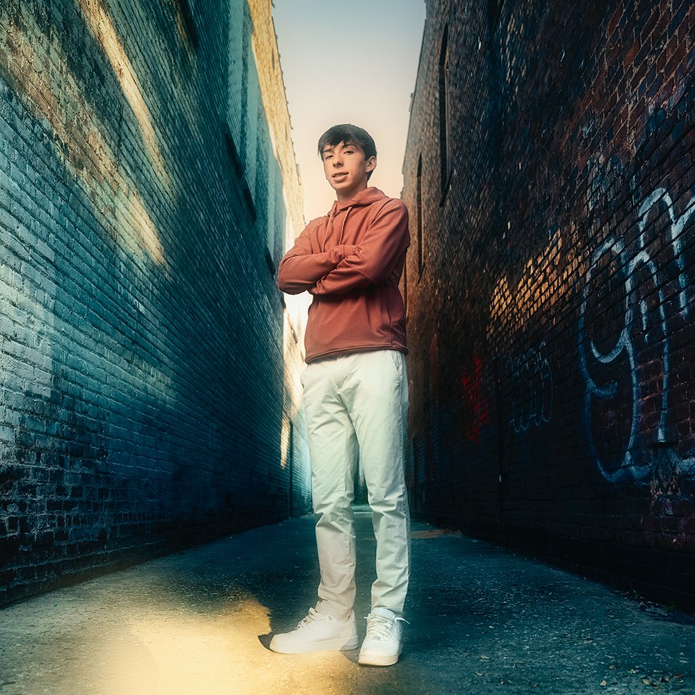 Don Evans Photography in Greensboro high school senior wearing khakis and a hoodie poses with hands on hips in an alley during his outdoor senior pictures photo session