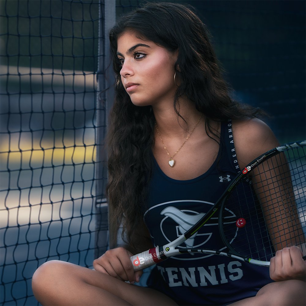 Don Evans Photography in Greensboro image of a Senior High School Tennis player sitting on the court looking at through the net during her senior banner photo session.jpg