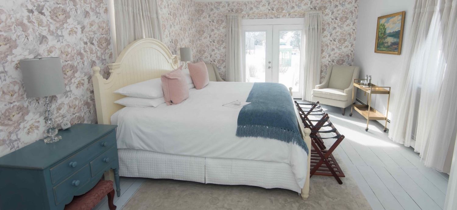 A bedroom with a large white bed, pink pillows, blue throw blanket, two bedside lamps, a blue dresser, a white armchair, a painting on the wall, a gold side table with glasses, and a wooden luggage rack near glass doors with curtains, letting in natural light.