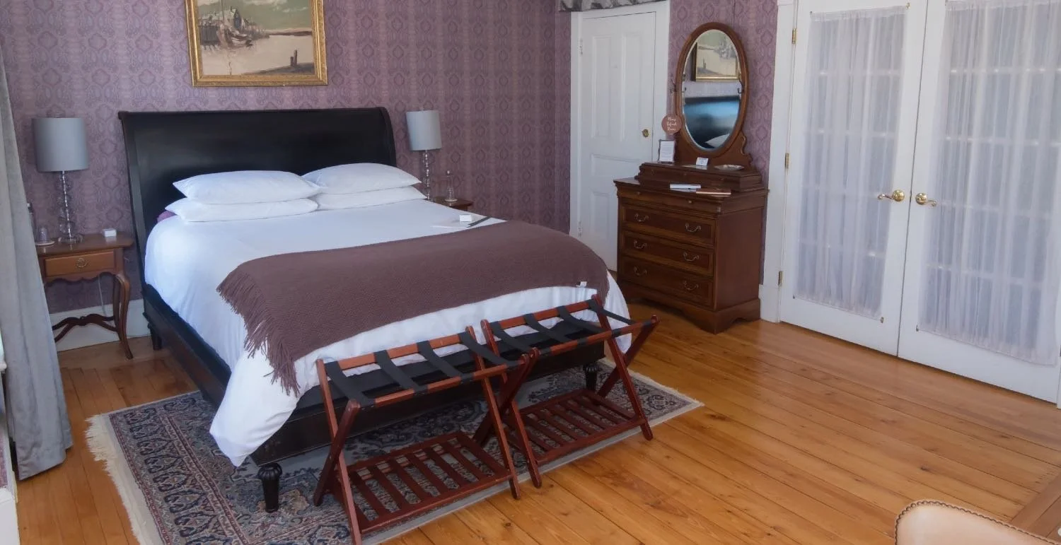 A bedroom with a large bed and bedding, a wooden dresser with a mirror, two nightstands with lamps, a painting on the purple patterned wallpaper, and wooden folding luggage racks in front of the bed.