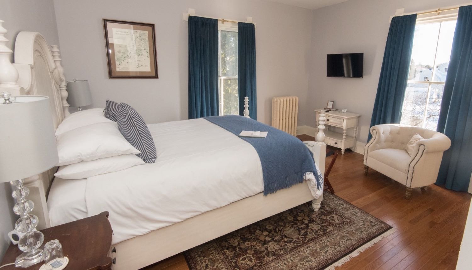 A neatly made bedroom with white bedding, blue throw, and striped pillows, featuring a white bed frame, blue curtains, a beige armchair, a small white nightstand, and a wall-mounted TV.