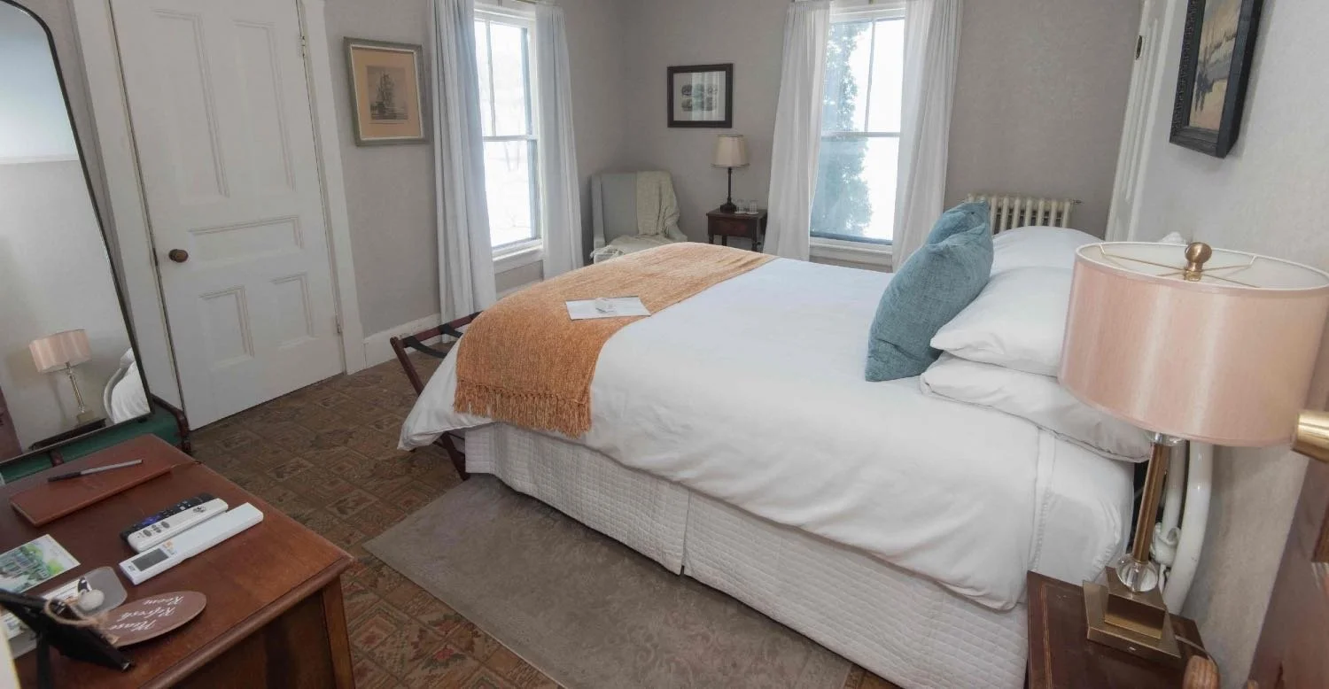 A cozy bedroom with a bed, a side table with a lamp, a closet, two windows with white curtains, a chair, and framed artwork on light-colored walls.