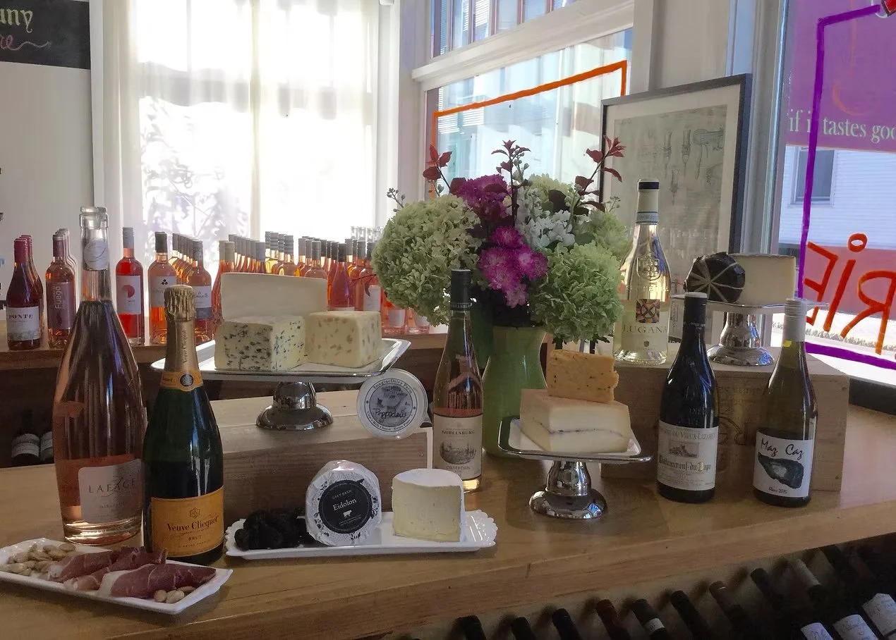 Cheese, wine bottles, and flowers displayed on a wooden counter in a bright room, with bottles lined up by a window.