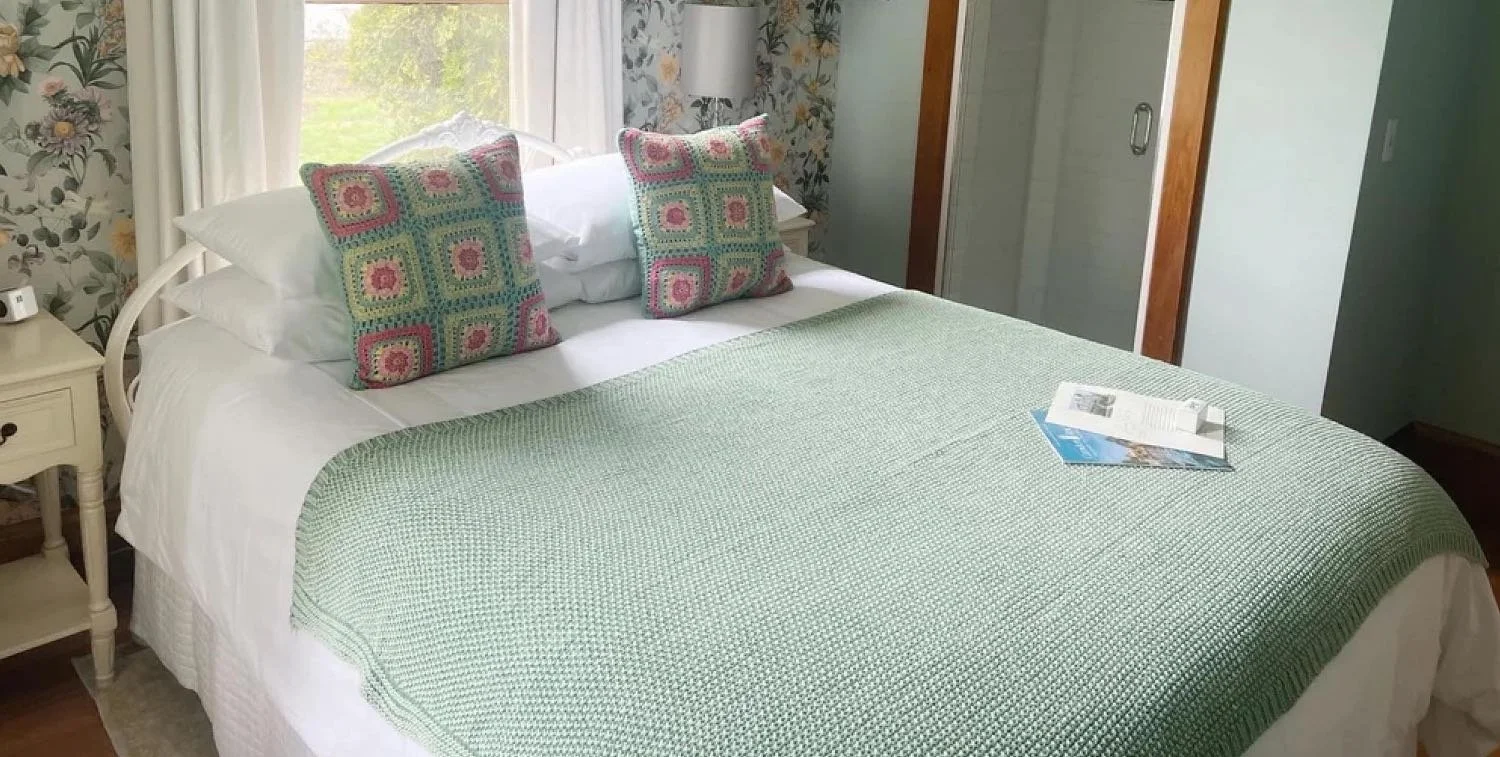 A neatly made bed with a green cover and two crocheted pillows with pink and green designs in a cozy bedroom with floral wallpaper, a window, and a nightstand.