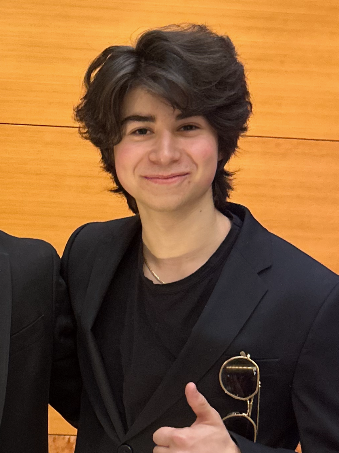 Young man with dark, wavy hair smiling and giving a thumbs up, wearing a black blazer and sunglasses hanging from the blazer pocket, standing against a wooden background.